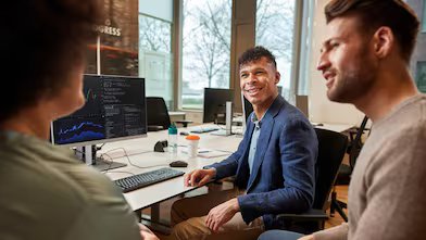 Three diverse people conversing in an office, with a computer monitor displaying graphs and codes in front of them. A man with dark curly hair in a blue blazer smiles at an out-of-focus woman with dark hair, while another man looks at her and appears to be engaged.