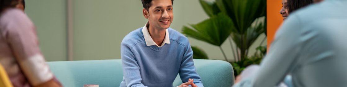 A person in a light blue sweater is seated in a lounge area, listening attentively to others. The setting includes colorful chairs, coffee mugs, and a relaxed atmosphere suitable for brainstorming or informal meetings.​