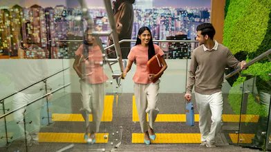 Two people walking down a modern indoor staircase with glass railings. One person is holding a folder, and the other is carrying a water bottle. Both are holding the handrails. The background features a large cityscape mural and a green wall decoration.”