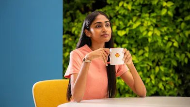 A person is holding a white coffee mug with the Shell logo, seated at a round table. The background features lush greenery, creating a refreshing and relaxed atmosphere.