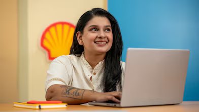 A person is working on a laptop at a desk with a Shell logo visible on the wall behind. A yellow notebook and pen are placed on the desk, suggesting a focused work environment. The background features vibrant blue and yellow panels.​