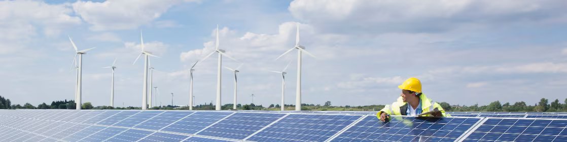 Male wearing a hardhat leaning on solar pannels with wind turbines in the background